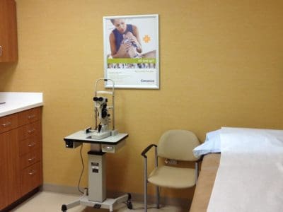 A clean medical examination room with an exam table, a beige chair, medical equipment, wooden cabinets, and a poster on a yellow wall.
