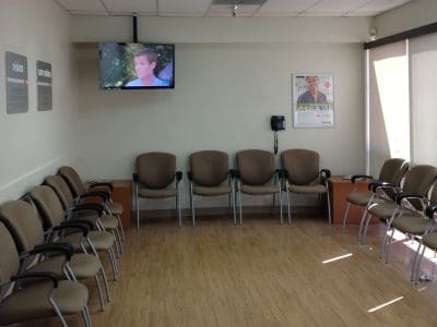 A waiting room with empty chairs arranged in a U-shape, a wall-mounted TV displaying a person, wall posters, and a phone on the wall. The room has wooden flooring and bright lighting.