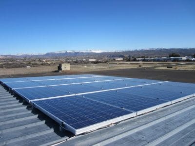 Solar panels from the City of Cottonwood Airport PV System are installed on a metal rooftop, with a clear sky and mountains visible in the background.