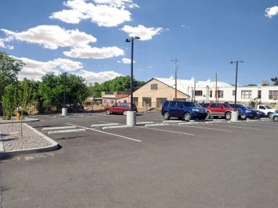 A mostly empty parking lot with a few cars parked, surrounded by trees and buildings under a blue sky with scattered clouds.