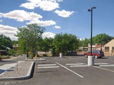 A mostly empty parking lot with a few parked vehicles, trees, a light pole, and buildings in the background under a partly cloudy sky.
