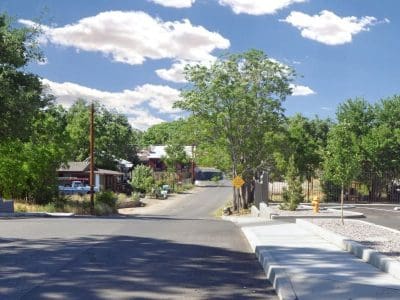 A quiet residential street with houses, trees, and parked cars under a blue sky with scattered clouds. The road curves slightly and a sidewalk lines the right side.
