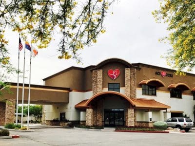 A two-story medical building with a red broken heart logo and the words “Acute Care” on the facade, located next to three flagpoles and surrounded by trees and parked vehicles.