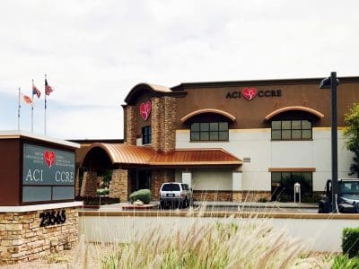 Medical office building with ACI and CCRE signage, stone accents, three flagpoles, and a parked vehicle in front. ACI CCRE logos feature red hearts.