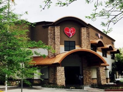 A two-story building with a large stylized heart logo above the entrance, surrounded by trees and a parking area in front.