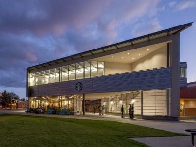 Modern two-story building with large windows at dusk, featuring a Starbucks on the ground floor, with people near the entrance and a cloudy sky overhead.