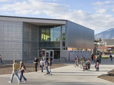 People walk, cycle, and skateboard near a modern glass and metal building on a campus with mountains visible in the background.