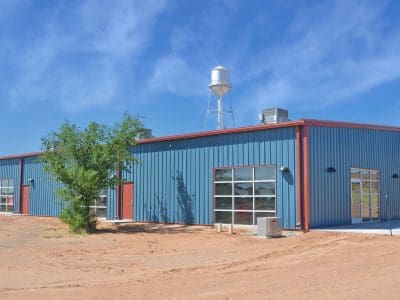A blue metal industrial building with large glass doors sits on a dirt lot under a clear sky, with a water tower visible in the background.