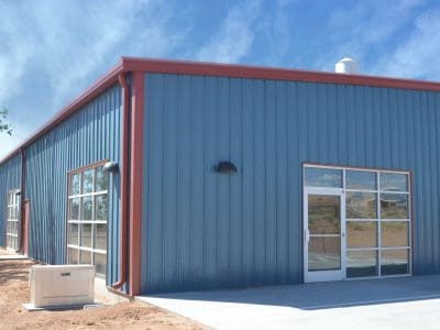 A modern blue metal building with large glass windows and doors, a red trim roof, and outdoor lights, under a clear blue sky.