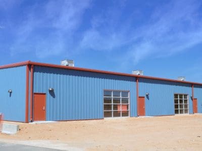 A large blue metal industrial building with red doors and multiple garage-style windows, set on a sandy lot under a clear blue sky.