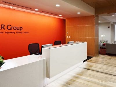 Modern office reception area with a white desk, two chairs, and DLR Group branding on an orange wall; seating area and stairs visible in the background.