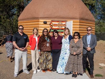 Seven people stand in front of a dome-shaped building with wooden siding and a brown roof, posing for a group photo outdoors on a sunny day.