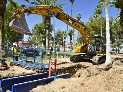 A construction excavator moves dirt near workers installing or inspecting a blue utility trench in a park with palm trees.