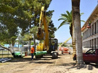 Construction workers operate heavy machinery and handle green pipes near a building and palm trees on a sunny day.