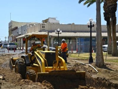 A construction crew operates a yellow backhoe digging a trench along a street with shops and palm trees in the background.