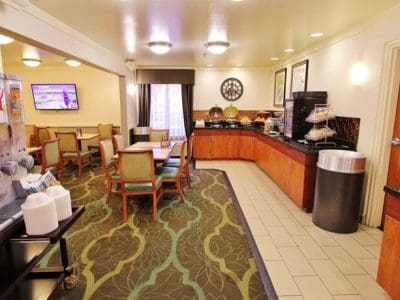 Breakfast area in a hotel with a cereal dispenser, tables and chairs, a buffet counter with food and beverages, and a TV on the wall.