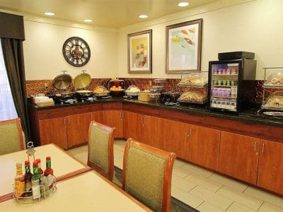 A hotel breakfast buffet with chafing dishes, fruit, pastries, a small fridge with drinks, and condiments on a table in the foreground.