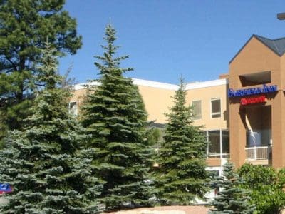A tan and white building partially obscured by pine trees, with a sign reading "Emergency Room Entrance" above the entrance.