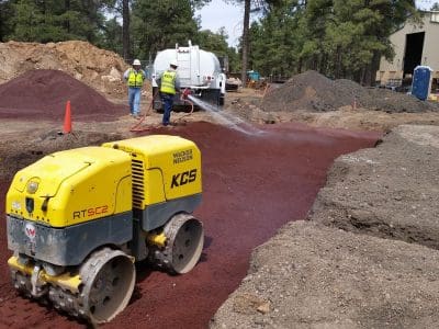 Construction workers spray water on a reddish dirt surface while a yellow compactor machine smooths the ground at a construction site surrounded by trees.