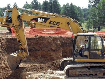 A person operates a yellow CAT excavator, digging a large trench at a construction site with orange safety fencing and trees in the background.