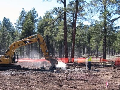 An excavator digs in a forested area while a construction worker in a safety vest observes nearby; orange safety fencing surrounds the site.
