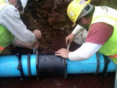 Two construction workers in safety gear secure a connector onto a large blue underground pipe at a work site.