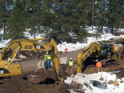 Construction workers and excavators are digging along a snow-lined roadside, with traffic signs and equipment visible in the background near a forested area.