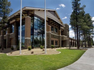 A modern two-story building with large glass windows, brick and stone exterior, surrounded by trees, grass, and a sidewalk under a blue sky.