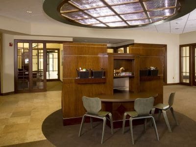 Modern office lobby with a semi-circular wooden desk, three chairs, overhead lighting, and glass doors leading to adjacent rooms.