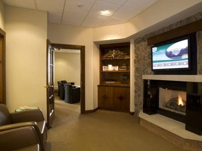 Waiting room with leather chairs, a fireplace below a wall-mounted TV, built-in shelves, and an open door leading to another room with more chairs.