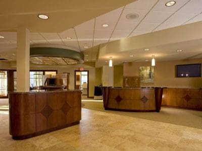 Spacious, well-lit office reception area with wooden desks, tiled floor, overhead lighting, a wall-mounted TV, and artwork on the wall.