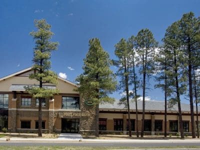 A large, two-story brick and stone building with tall windows, surrounded by pine trees, under a clear blue sky.