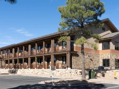 Two-story building with stone and wood exterior, outdoor staircases, and balconies, surrounded by trees and a parking lot under a clear sky.