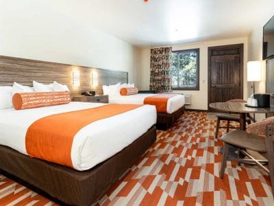 Hotel room with two double beds, orange and white bedding, patterned carpet, a table with two chairs, a TV, dresser, and a window with curtains.