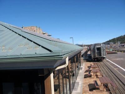 Outdoor seating area next to the long building with a green roof, featuring the Xanterra Grand Depot Café PV System, beside train tracks where an old-fashioned railcar is parked under a clear blue sky.