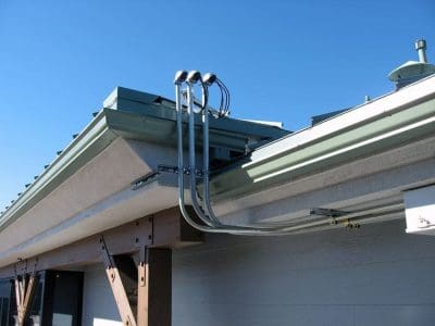 Four metal electrical conduits run up the exterior wall and into the roofline of a building, part of the Xanterra Grand Depot Café PV System, with a blue sky in the background.