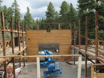 Partially constructed wooden building frame with exposed beams and a single completed wall; construction equipment is present in the center. Pine trees are visible in the background.