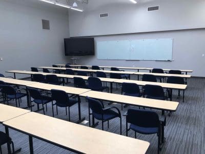 Empty classroom with rows of desks and chairs, a large whiteboard on the wall, and a flat-screen monitor in the front. The room has gray walls and a carpeted floor.