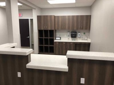 Reception area of an office or clinic with wood cabinetry, open cubby shelves, countertop workspace, and a sink. Walls and counters are neutral tones, and fluorescent lighting is overhead.