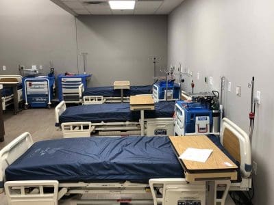 A hospital room with several empty adjustable beds, side tables, medical equipment, and IV stands arranged in a neat, orderly manner.