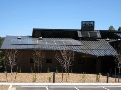 A building with a metal roof features the Highlands Fire District PV System installed on top, young trees and a mulched area in the foreground, and a clear blue sky above.
