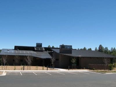 A modern, single-story building with a dark metal roof and solar panels from the Highlands Fire District PV System, set against a clear blue sky, with an empty parking lot in the foreground.