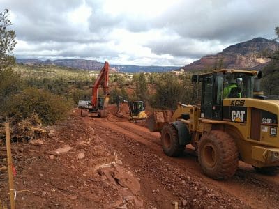 Construction vehicles, including a loader and excavator, work on a dirt road in a rocky, forested landscape with mountains in the background.