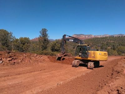 A yellow excavator is moving dirt on a cleared construction site, surrounded by trees and mountains under a clear blue sky.