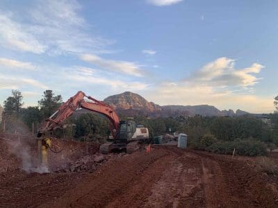 An excavator breaks up rocky ground on a dirt construction site with mountains and scattered clouds visible in the background.