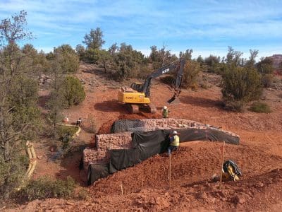 A construction site in a desert area shows workers and an excavator building a rock-filled retaining wall with a black protective barrier.