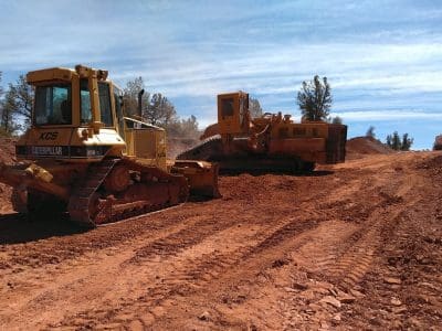 Two yellow Caterpillar bulldozers move and level dirt on a construction site under a partly cloudy sky, with trees in the background.