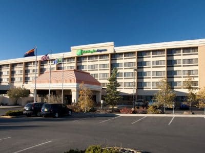 A four-story Holiday Inn Express hotel with a parking lot in front, several cars parked, and multiple flags displayed near the entrance on a clear day.