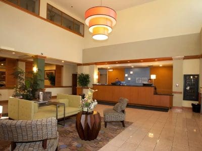 Hotel lobby with modern seating area, patterned chairs, central table with flowers, and reception desk in the background illuminated by a large ceiling light fixture.