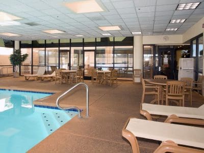 Indoor swimming pool area with lounge chairs, tables, and chairs on a tiled floor, surrounded by large windows and glass doors.